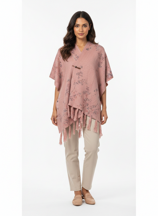 Model wearing a Cozy&Drape dusty pink floral poncho wrap with a button closure and fringe hem over beige trousers.